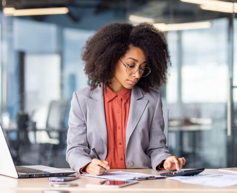 Attentive financial manager checking accuracy of counting on calculator and analyzing bills laying on desktop with laptop. Concentrated female in suit managing expenses of company in modern office.