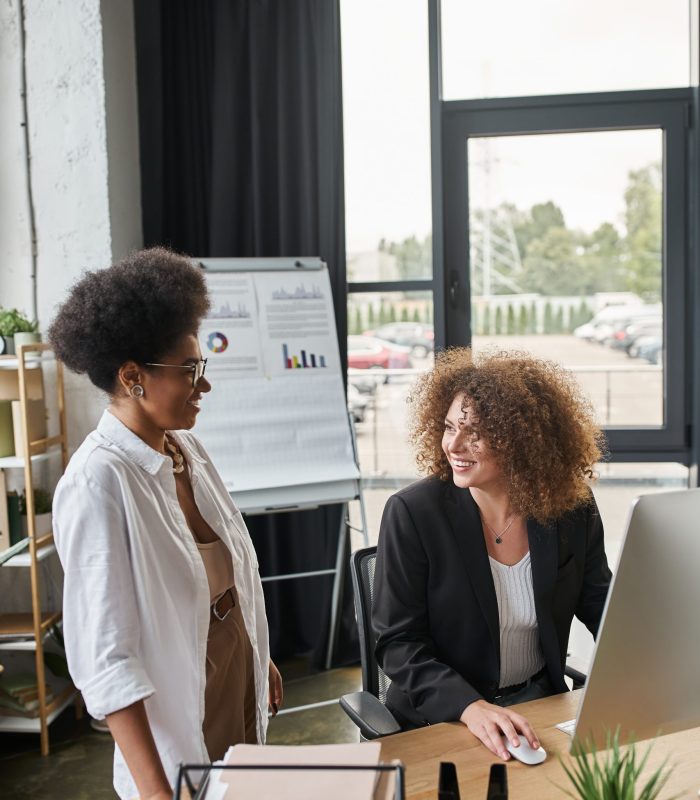 smiling african american businesswoman near female colleague working on computer in office, teamwork