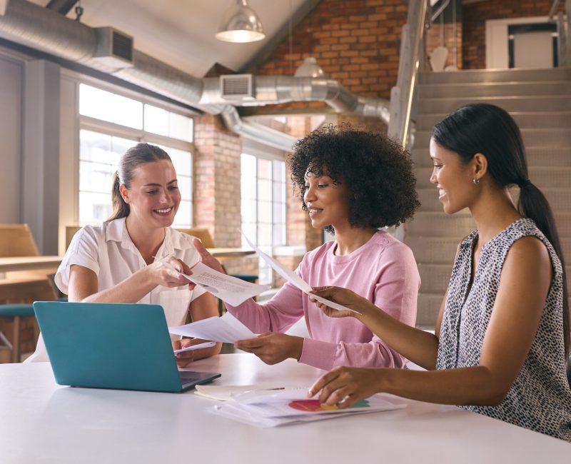 Three Young Businesswomen Meeting In Modern Office Sitting Around Table Working On Laptop Together